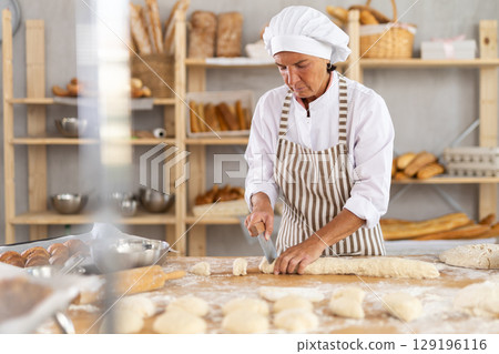 Mature woman baker standing at table in bakehouse and cutting dough into pieces. Mature woman baker standing at table in bakehouse and cutting dough into pieces. 129196116