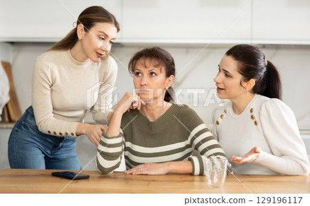 Sad woman sits silently in kitchen, adult girls sympathize and comfort her Sad woman sits silently in kitchen, adult girls sympathize and comfort her 129196117