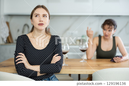 Upset middle-aged woman sitting at the kitchen table with her back to another lady quarreling to her Upset middle-aged woman sitting at the kitchen table with her back to another lady quarreling to her 129196136