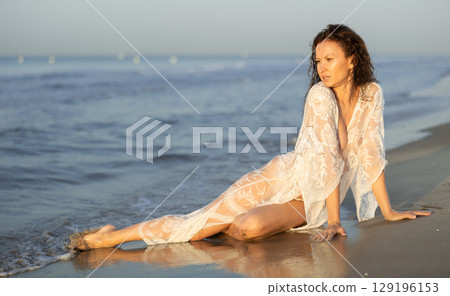 Adult woman in white peignoir sitting on coast Adult woman in white peignoir sitting on coast 129196153
