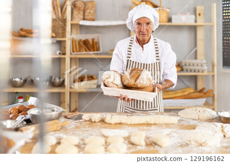 Polite mature female baker with fresh bread posing in bakery Polite mature female baker with fresh bread posing in bakery 129196162