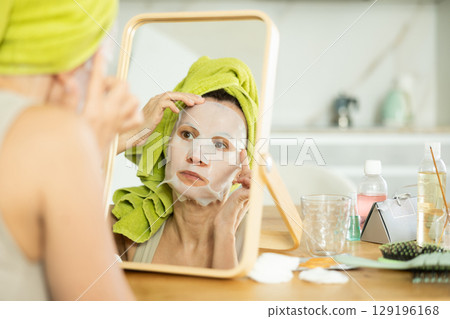 Middle-aged woman applying tissue mask on her facial skin sitting in front of the mirror Middle-aged woman applying tissue mask on her facial skin sitting in front of the mirror 129196168