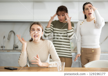 Upset middle-aged woman sitting at the kitchen table with her back to women quarreling to her Upset middle-aged woman sitting at the kitchen table with her back to women quarreling to her 129196177