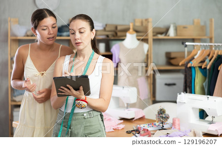 Woman tailor and girl client discuss manufacture of clothes, look at tablet screen Woman tailor and girl client discuss manufacture of clothes, look at tablet screen 129196207