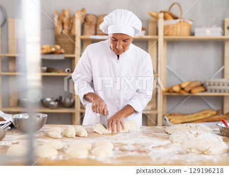 Confidence elderly female baker in white uniform standing at work table and cutting dough into pieces in kitchen of bakery 129196218