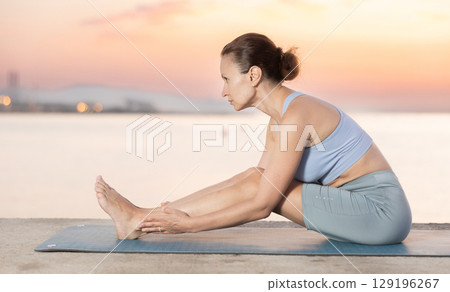 Concentrated young woman making stretching legs sitting on yoga mat and doing exercise yoga on beach at sunrise 129196267