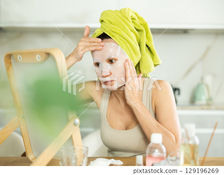Woman applying moisturizing sheet face mask in front of mirror 129196269