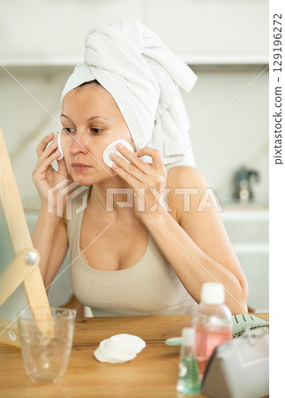 Middle-aged woman cleaning her facial skin with cotton disks sitting in front of the mirror Middle-aged woman cleaning her facial skin with cotton disks sitting in front of the mirror 129196272