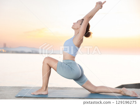 Skilled happy young female teacher demonstrating variation of warrior pose of yoga on seashore at sunrise Skilled happy young female teacher demonstrating variation of warrior pose of yoga on seashore at sunrise 129196275