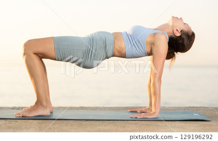 Portrait of caucasian experienced woman yoga teacher performs ardha chakrasana on seashore at sunset Portrait of caucasian experienced woman yoga teacher performs ardha chakrasana on seashore at sunset 129196292