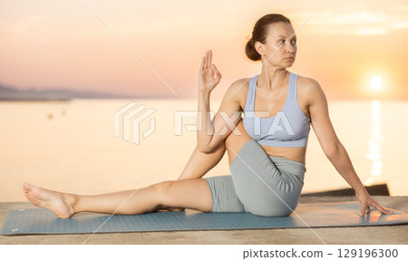 Calm european young female yoga trainer practicing Half Lord of fishes pose or Ardha Matsyendrasana during yoga training on seashore at sunrise Calm european young female yoga trainer practicing Half Lord of fishes pose or Ardha Matsyendrasana during yoga training on seashore at sunrise 129196300