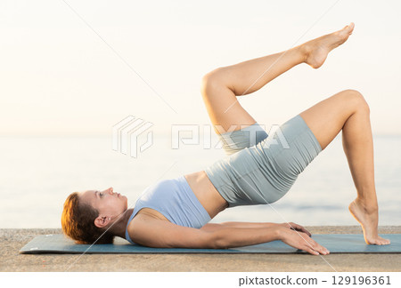 Sporty female wearing sportswear standing legs up in Salamba Sarvangasana exercise during yoga on seashore at sunrise Sporty female wearing sportswear standing legs up in Salamba Sarvangasana exercise during yoga on seashore at sunrise 129196361