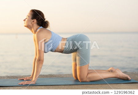 Skilled young female trainer performing cat-cow pose of yoga on seashore at sunrise Skilled young female trainer performing cat-cow pose of yoga on seashore at sunrise 129196362