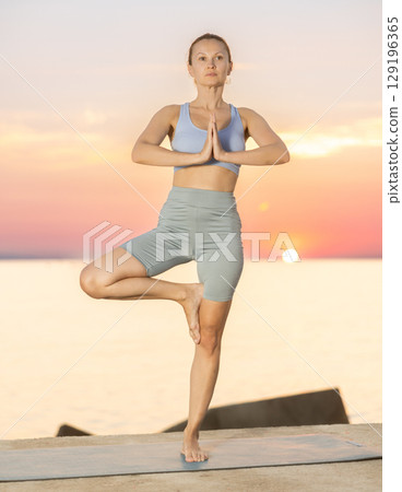 Sedulous woman doing warrior one leg pose of yoga on mat on beach at sunrise Sedulous woman doing warrior one leg pose of yoga on mat on beach at sunrise 129196365