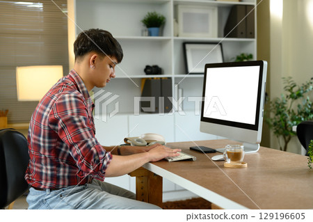 Side view of man working from home on desktop computer with coffee cup on wooden desk 129196605