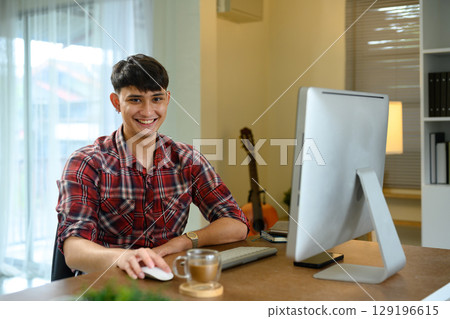 Young man smiling while working on a desktop computer at home office desk 129196615
