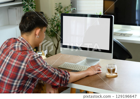 Side view of man working from home on desktop computer with coffee cup on wooden desk Side view of man working from home on desktop computer with coffee cup on wooden desk 129196647