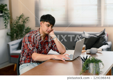 Young man working on a laptop at home, appearing thoughtful and slightly bored Young man working on a laptop at home, appearing thoughtful and slightly bored 129196648