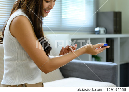 Smiling young woman applying moisturizing cream on her arm while sitting on a sofa at home 129196666
