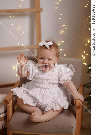 Smiling baby girl in white dress sitting on chair and waving during Christmas at home with lights 129196688