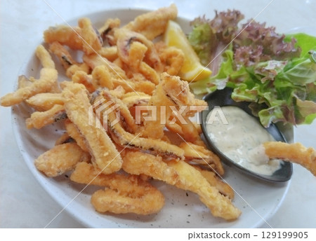Golden, crispy fried squid calamari rings served on a dark slate plate with zesty lemon wedges and creamy garlic aioli. 129199905
