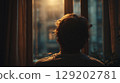 Back view of a man gazing out a window, illuminated by warm light. 129202781