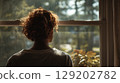 Rear view of a young woman with curly hair looking out a window quietly. 129202782