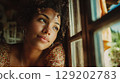 A contemplative young woman looking out her home window, daydreaming. 129202783