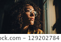 Close-up of a contemplative female in glasses by window in warm light. 129202784