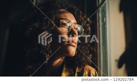 Close-up of a contemplative female in glasses by window in warm light. Close-up of a contemplative female in glasses by window in warm light. 129202784