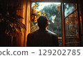Young man from behind, looking out open window at sunny garden plants and flowers. 129202785