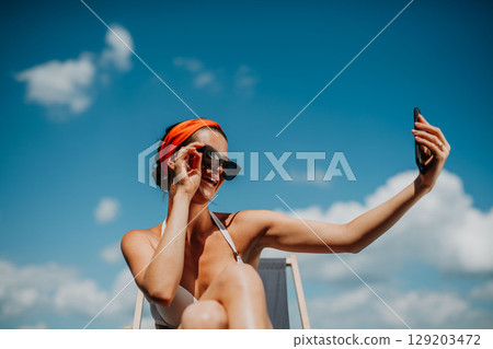 Young woman sitting by swimming pool outdoors in backyard garden, taking selfie. 129203472