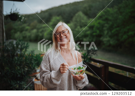 Senior woman standing terrace, eating healthy dinner outdoors. Senior woman standing terrace, eating healthy dinner outdoors. 129203512
