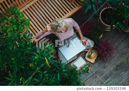 Older woman working from homeoffice, sitting on terrace with laptop. 129203596