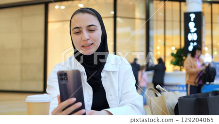 Young muslim woman in hijab relaxing at shopping mall table, browsing smartphone while enjoying takeaway coffee amid recent shopping bags, representing modern urban lifestyle 129204065