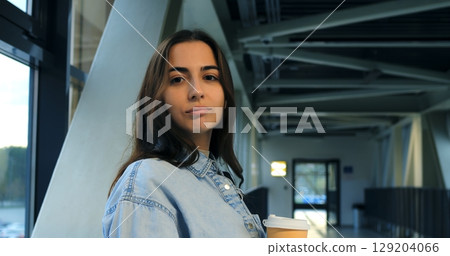 Stylish young woman drinking coffee, standing in modern building with large windows, experiencing peaceful moment of solitude and quiet reflection 129204066