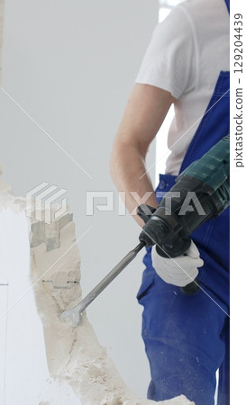 Unknown male construction worker wearing blue overalls is demolishing white wall with rotary hammer drill, generating dust, close up horizontal view. Renovation 129204439