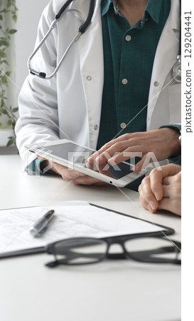 Doctor man in lab coat over a green shirt, and stethoscope is using tablet while consulting patient in modern medical office. Medicine and health care concept 129204441