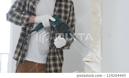Close up of unrecognizable male construction worker wearing beige checkered shirt and protective gloves, is demolishing white wall with rotary hammer drill, generating dust, close up view. Renovation 129204445