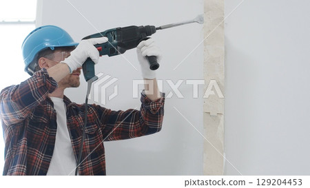 Male construction worker wearing red checkered shirt, blue protective helmet and gloves, is demolishing white wall with rotary hammer drill, generating dust, portrait view. Renovation concept 129204453