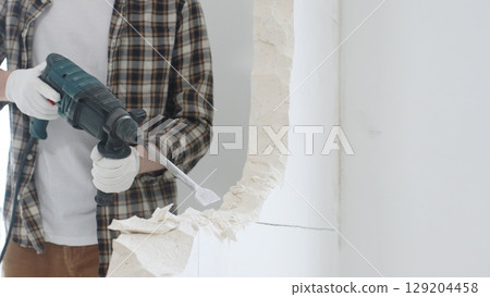 Close up of unrecognizable male construction worker wearing beige checkered shirt and protective gloves, is demolishing white wall with rotary hammer drill, generating dust, close up view. Renovation 129204458