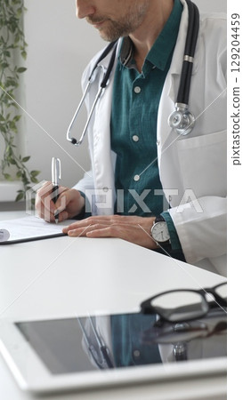 Doctor man wearing lab coat over green shirt, and stethoscope is writing medical prescription or filling medical documents sitting at office desk. Medicine and health care Doctor man wearing lab coat over green shirt, and stethoscope is writing medical prescription or filling medical documents sitting at office desk. Medicine and health care 129204459
