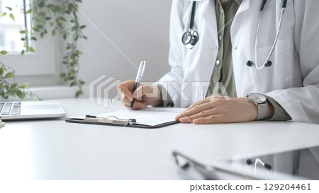 Professional doctor woman wearing white lab coat and stethoscope documenting patient information on clipboard near laptop in clinic workspace. Medicine and health care 129204461
