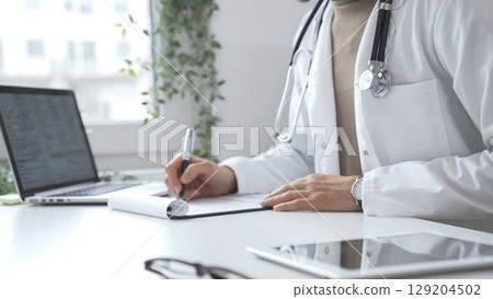 Male medical professional working at desk, writing prescription, wearing white lab coat and stethoscope in clinical workspace. Medicine and health care 129204502
