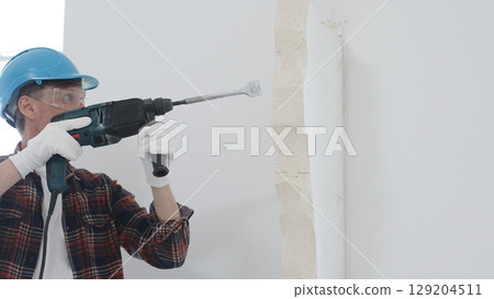 Man construction worker wearing red checkered shirt, blue hard hat and protective gloves, is demolishing white wall with rotary hammer drill, generating dust, portrait view. Renovation concept Man construction worker wearing red checkered shirt, blue hard hat and protective gloves, is demolishing white wall with rotary hammer drill, generating dust, portrait view. Renovation concept 129204511