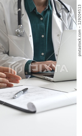 Doctor man wearing lab coat over a green shirt, is typing on a laptop and listening to a woman patient explaining symptoms during medical consultation in clinic. Medicine and health care concept Doctor man wearing lab coat over a green shirt, is typing on a laptop and listening to a woman patient explaining symptoms during medical consultation in clinic. Medicine and health care concept 129204513