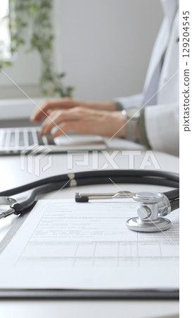 Stethoscope and medical records are on the desk opposite the doctor is typing on a laptop with a in a medical office, suggesting online healthcare or telemedicine services. Medicine concept Stethoscope and medical records are on the desk opposite the doctor is typing on a laptop with a in a medical office, suggesting online healthcare or telemedicine services. Medicine concept 129204545