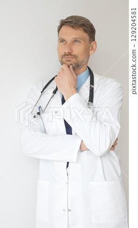 Portrait of a confident male doctor wearing stethoscope and white coat, standing with crossed arms and gazing thoughtfully, representing medical professionalism and expertise 129204581