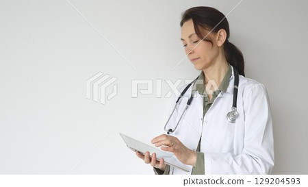 Headshot of a doctor woman wearing white medical coat over green blouse, stethoscope, is reviewing patient records on digital tablet, standing in medical workspace with natural lighting. Medicine 129204593