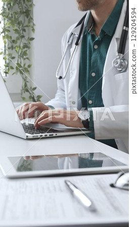 Doctor man wearing a white coat over green shirt, and stethoscope is typing on a laptop in a bright medical office, showcasing the intersection of healthcare and technology. Medicine and health care 129204594